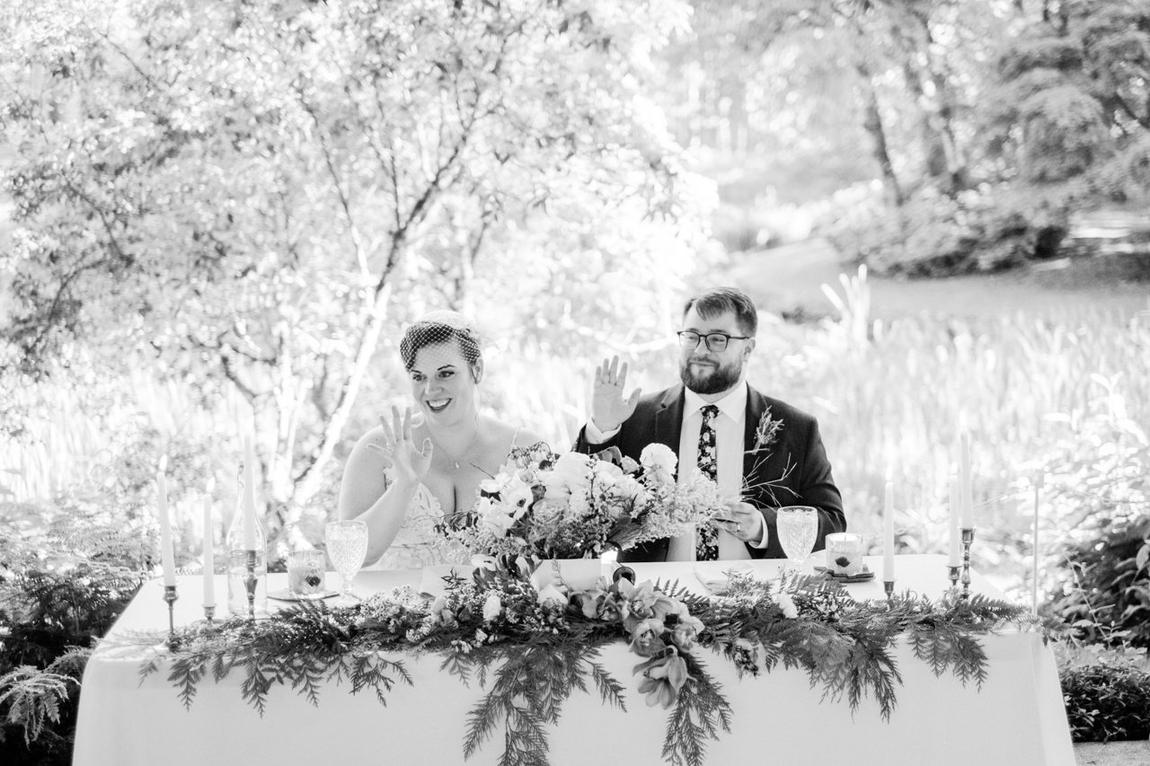  Bride and groom sit at head table decorated with ferns and wave to guests 