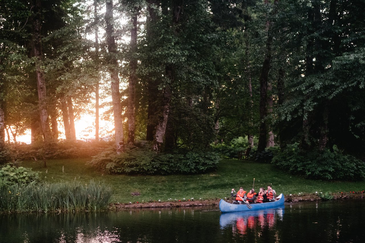  Wedding guests canoe in lake with orange glow of sunset behind them 