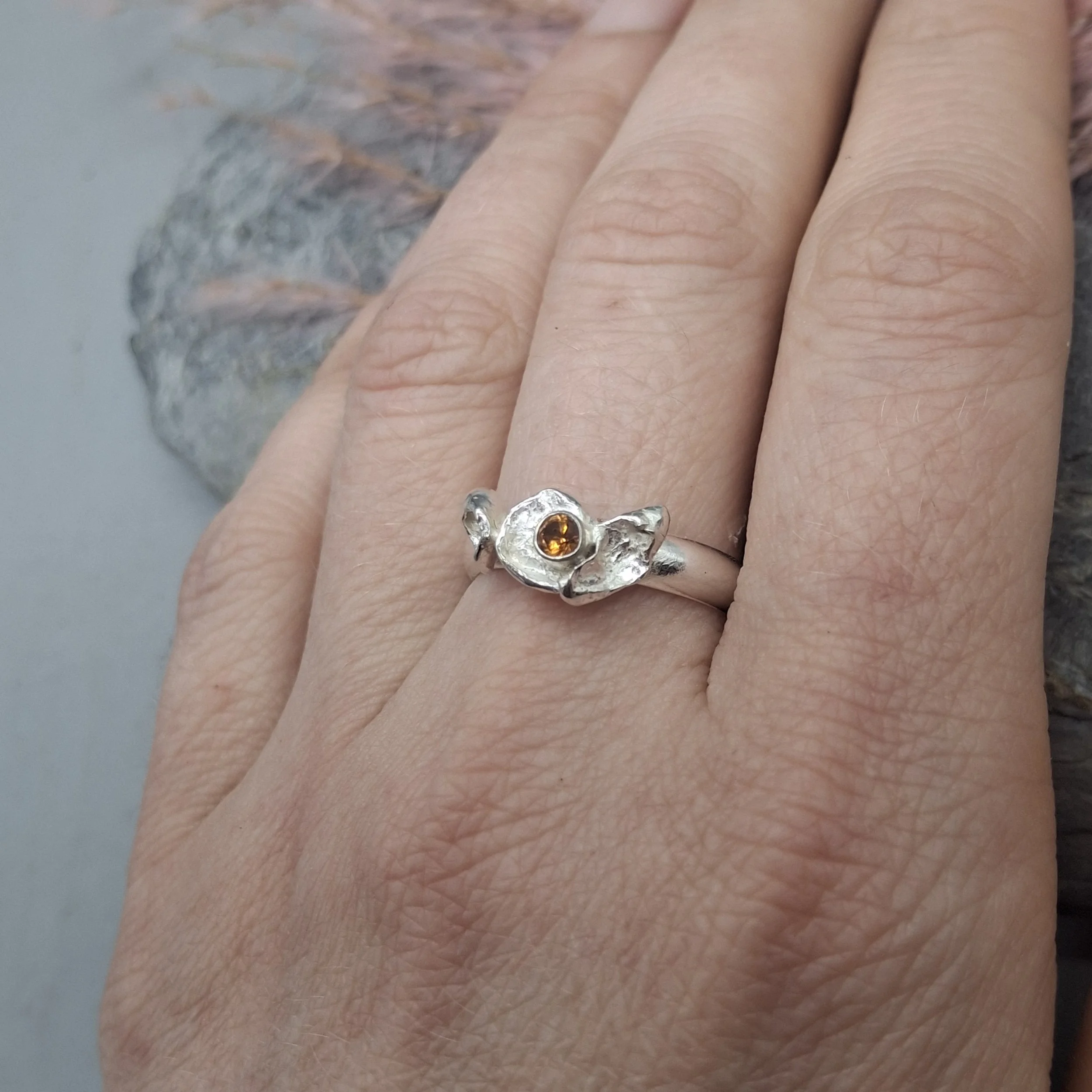 Handmade sterling silver Fallen Blossom ring with organic water-cast silver petals, featuring a glowing mandarin garnet in warm orange tones, shown worn on the hand