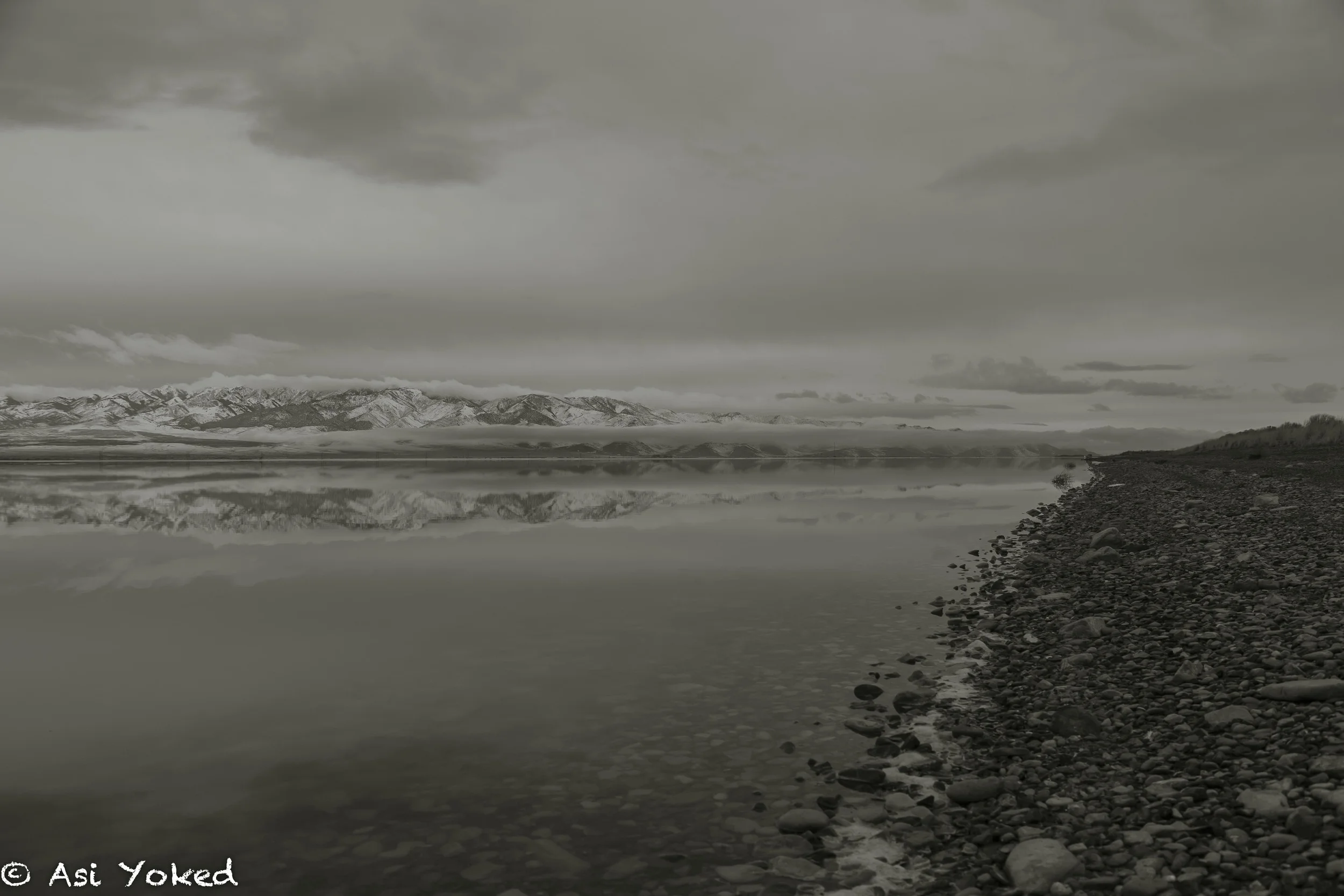 A calm lake with a rocky shoreline on the right side, reflecting snow-capped mountains under a cloudy, overcast sky.