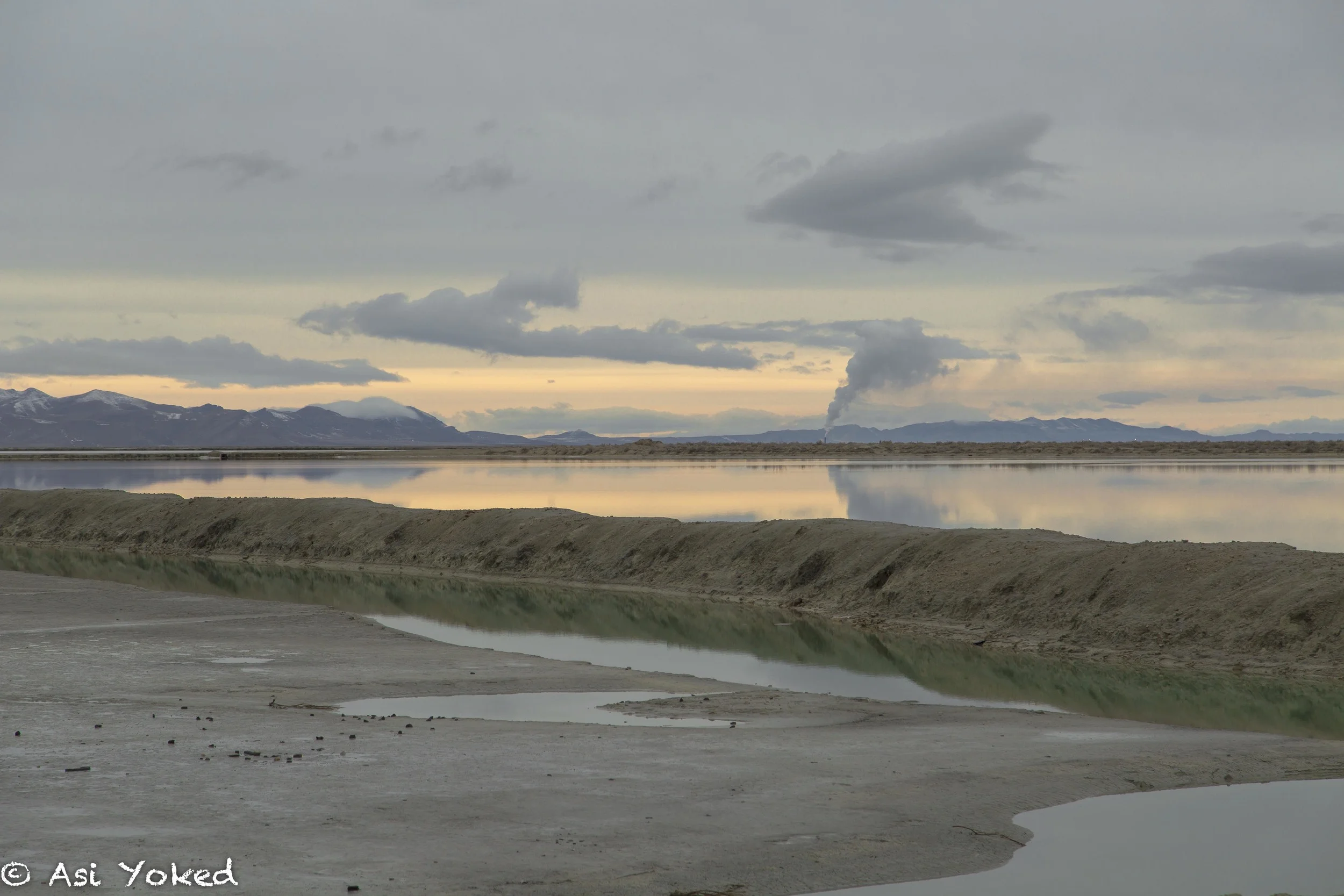 A landscape with a cloudy sky over mountains and a body of water reflecting the sky. Steam rises from a distant spot, possibly a geothermal feature, and the foreground shows a sandy, barren area with scattered rocks.