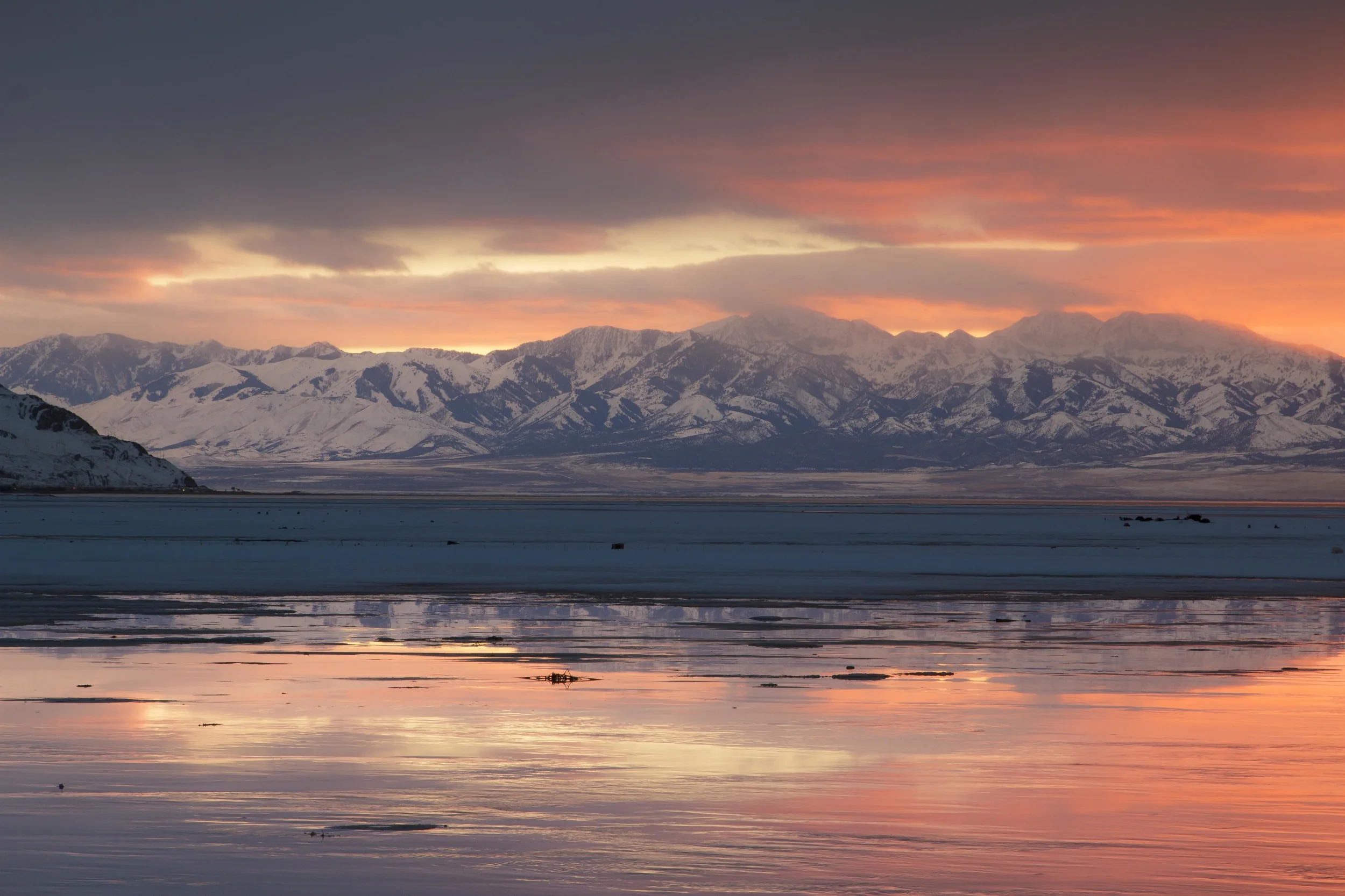 Snow-capped mountains at sunset with pink and orange clouds reflected in the water below.