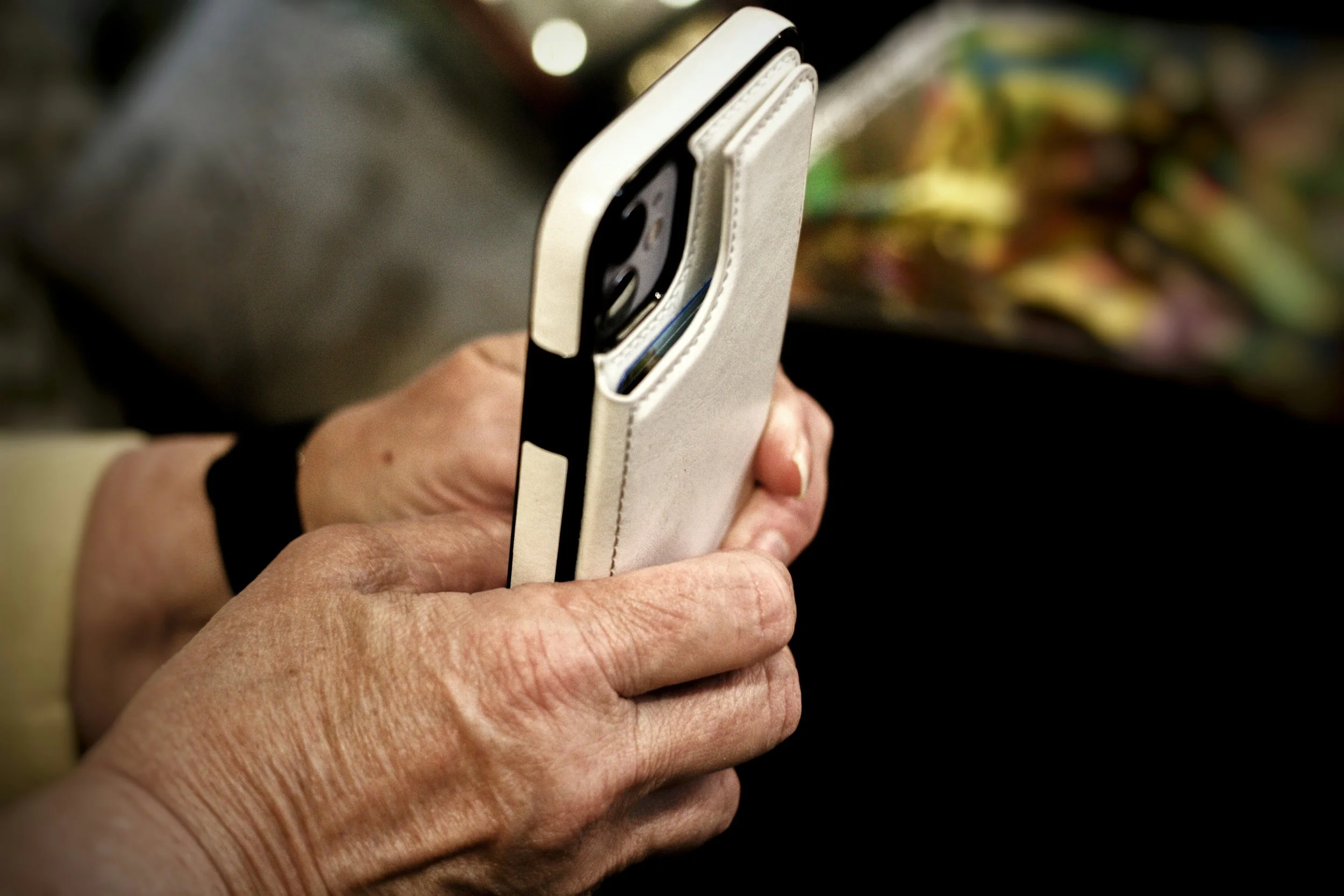 Close-up of an elderly person's hand holding a smartphone with a beige case, with a blurred colorful background.
