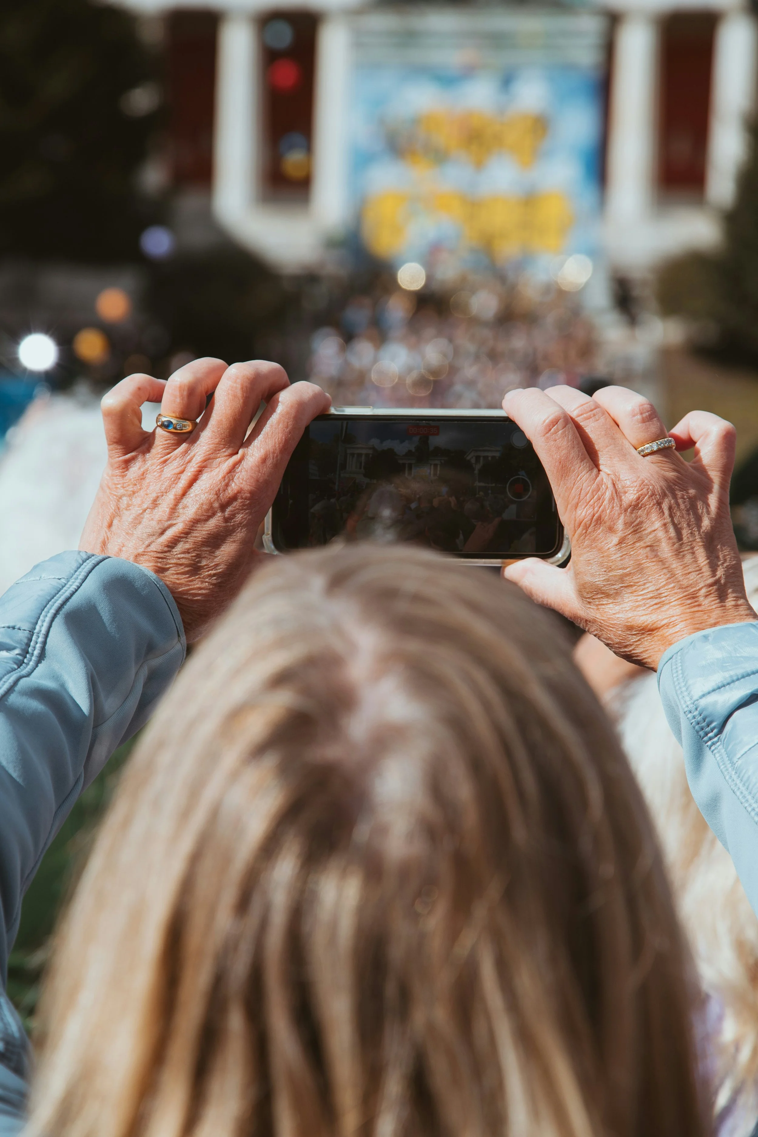 A person with light skin and blonde hair taking a photo of a float at a parade with a smartphone. The float has a blurred backdrop with bright colors and decorations. The person's hands are visible, wearing rings on both fingers. The person is wearing a light blue jacket.