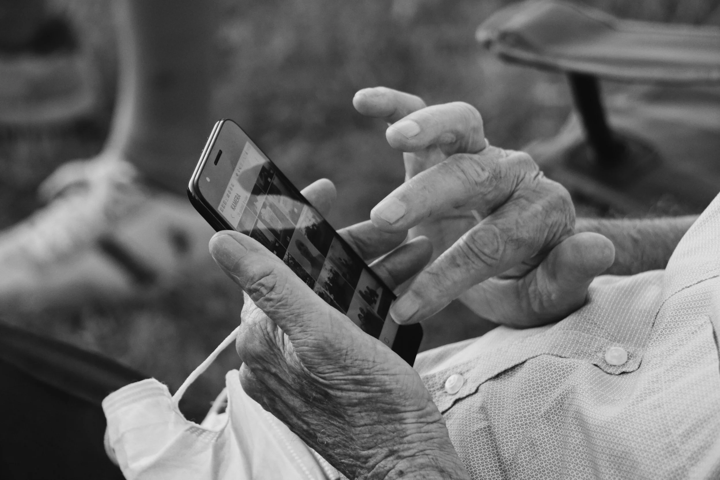 Close-up of elderly person's hand holding a smartphone, with another person's hand supporting it, both wearing long sleeves, in black and white.