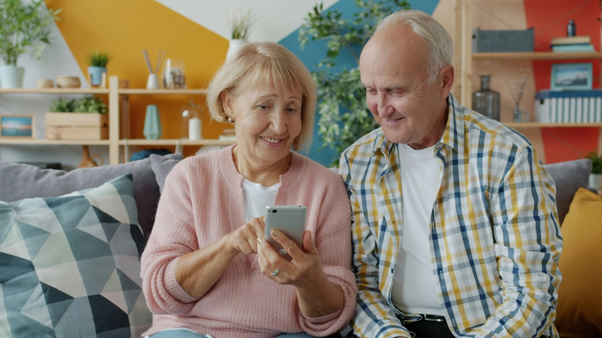 An elderly woman and man sitting on a couch, looking at a smartphone together in a colorful living room.