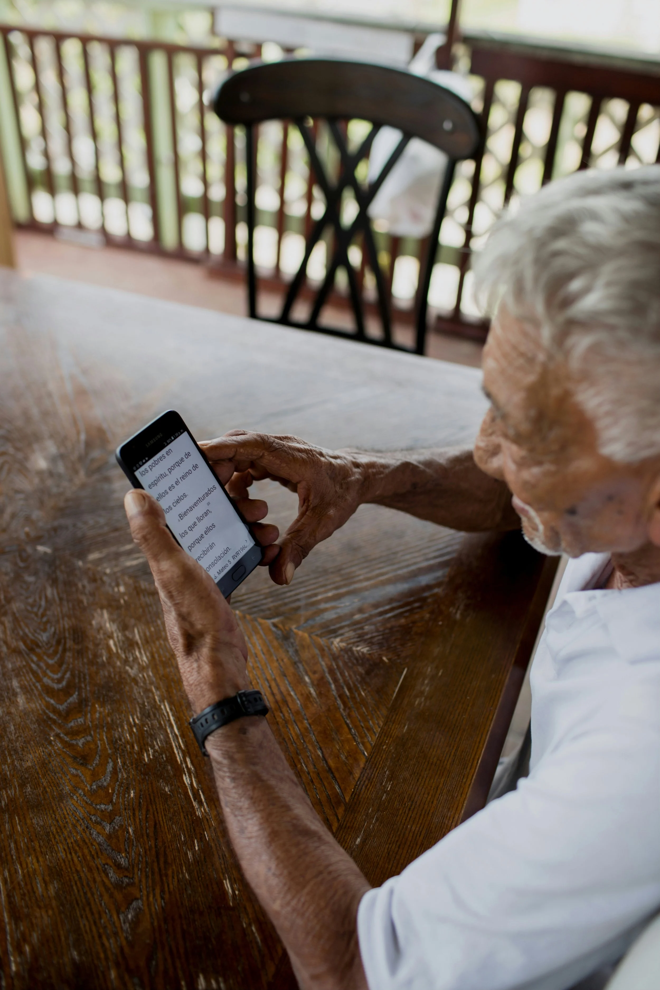 An elderly person with white hair sitting at a wooden table, holding a smartphone, with a blurred background of a balcony railing and greenery.