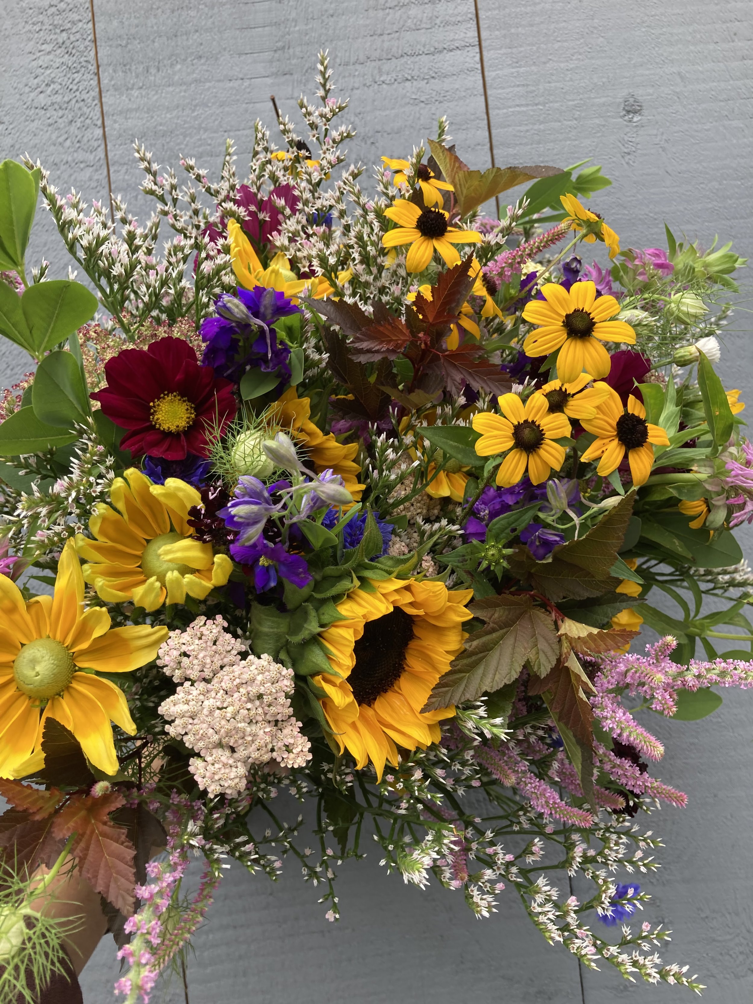 A handful of small market bouquets against a blue barn.  The bouquets contain yellow rudbeckia, sunflowers, pink rattail statice, yarrow, cosmos, german statice, ninebark, larkspur, and baptisia foliage.