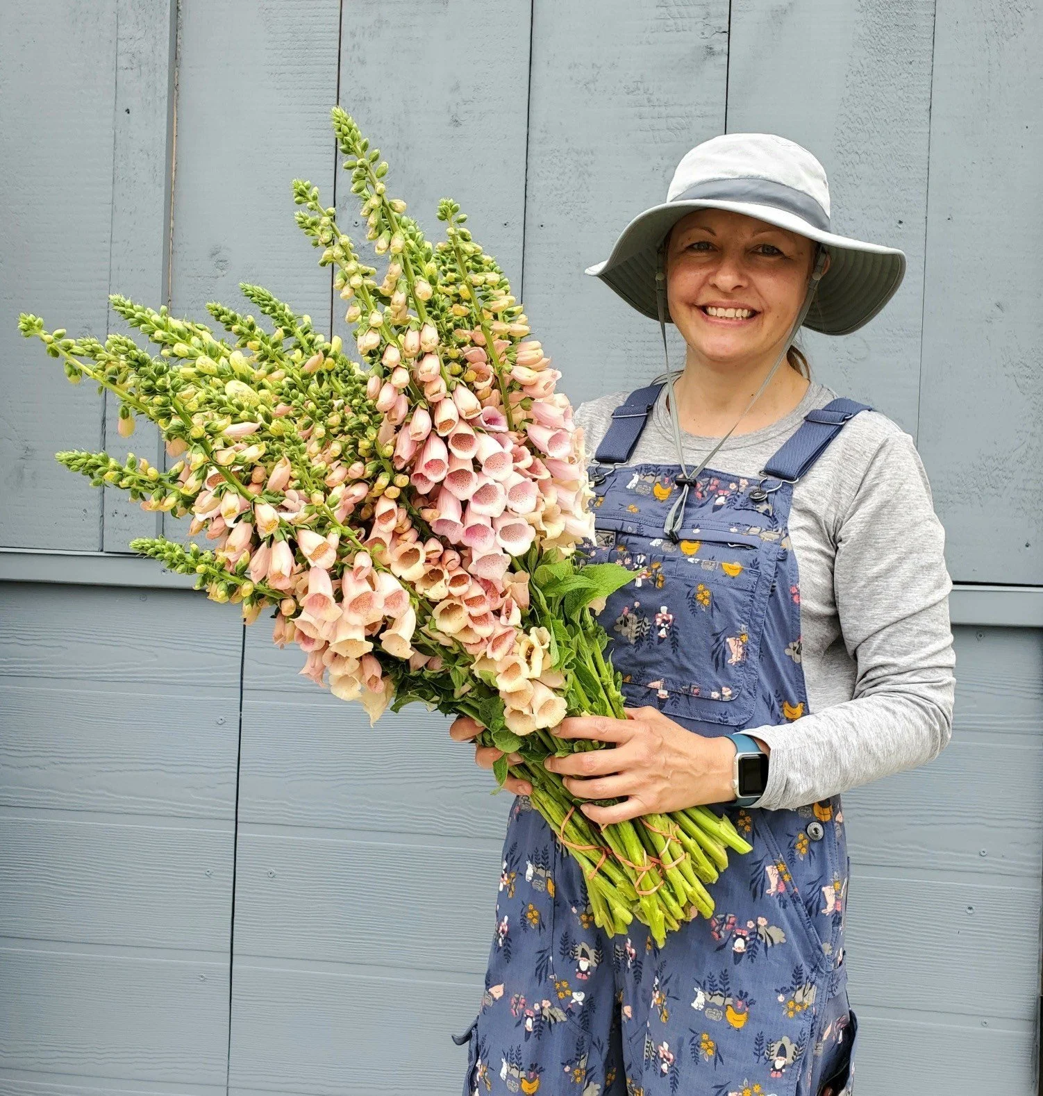 a white woman wearing a hat, blue overalls, and a grey shirt, is holding a large bundle of pink foxglove.