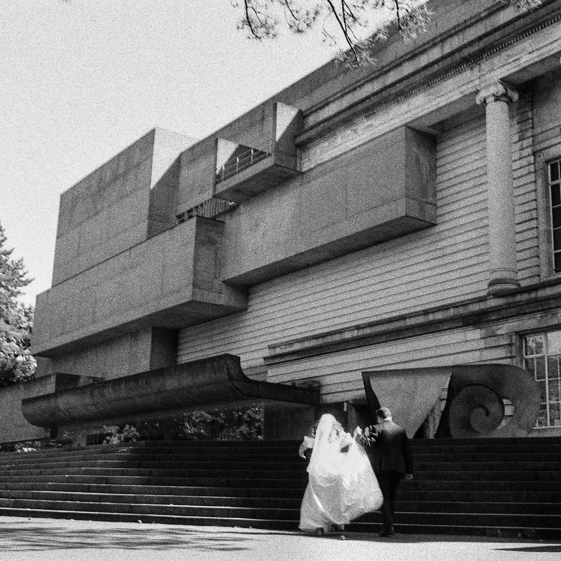 Wedding photo of married couple walking towards from facade of the ulster museum shot on analogue medium B&W film