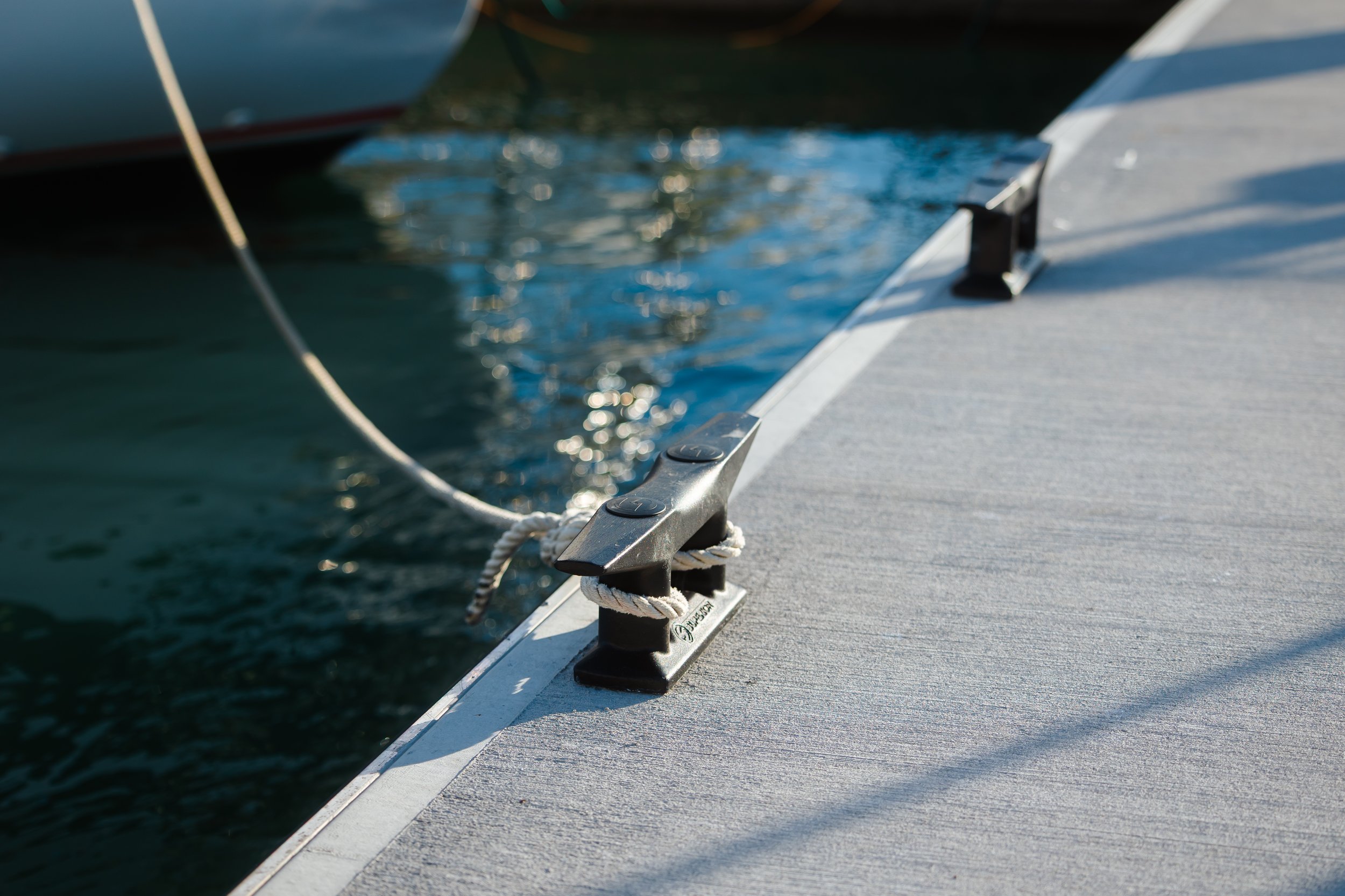 Close-up of boat cleats on a dock with a rope tied around it, water and boats in the background.