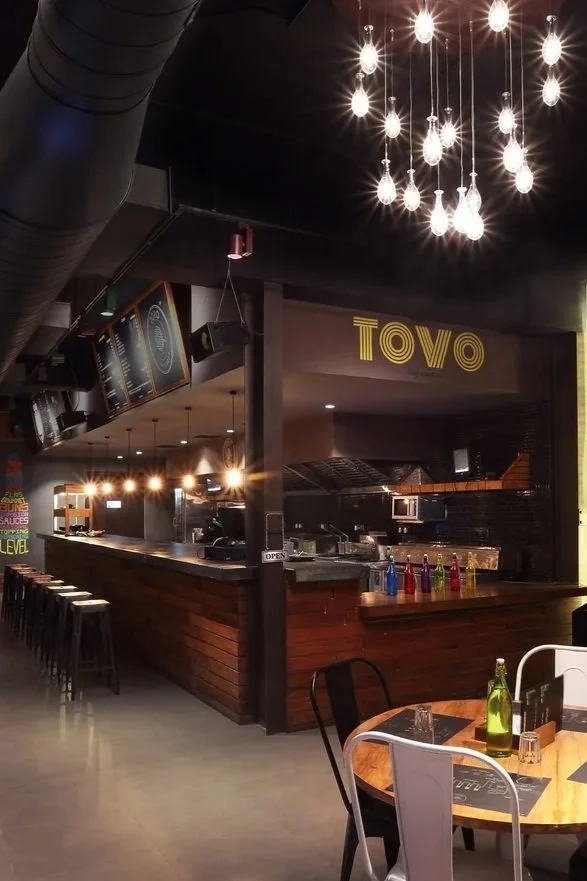 Interior of a modern restaurant with wooden tables, chairs, and an illuminated artwork on the wall featuring animated fish characters. The bar counter is visible with various colored bottles and an overhead sign reading 'Tovo.' Hanging lights illumin