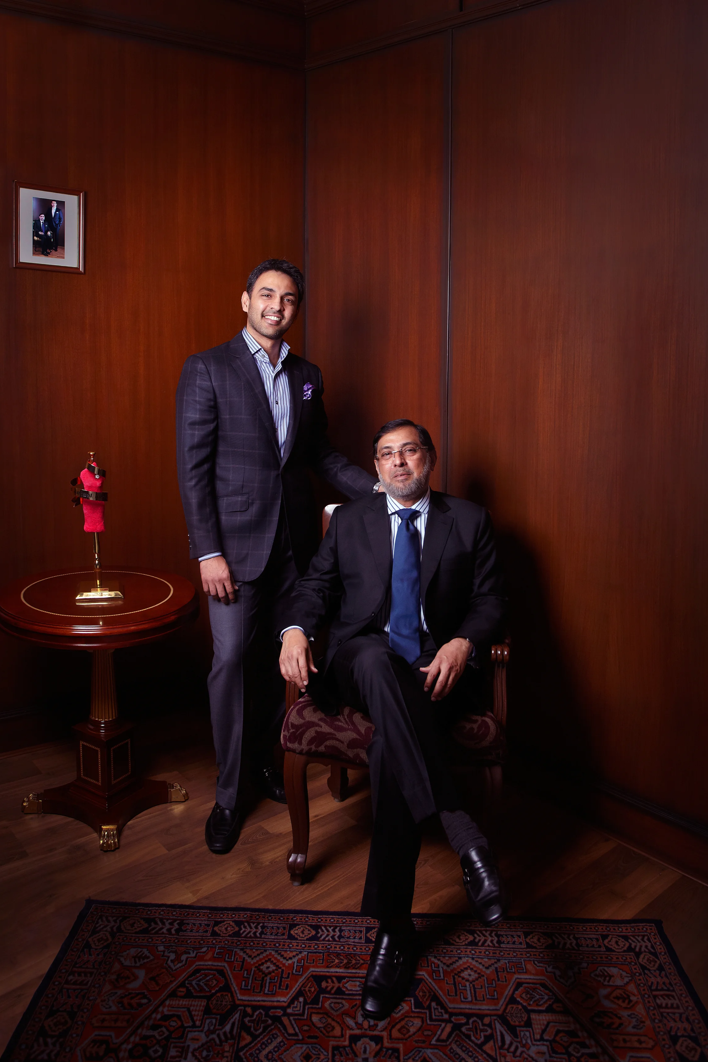 Two men in formal suits pose in a wood-paneled room, one sitting in a chair and the other standing beside him. A small table with a red statue and framed photo is nearby, along with an ornate carpet on the floor.