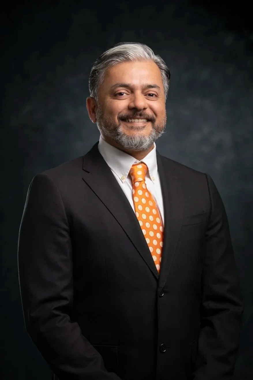Portrait of a man in a suit with an orange polka-dot tie against a dark background.