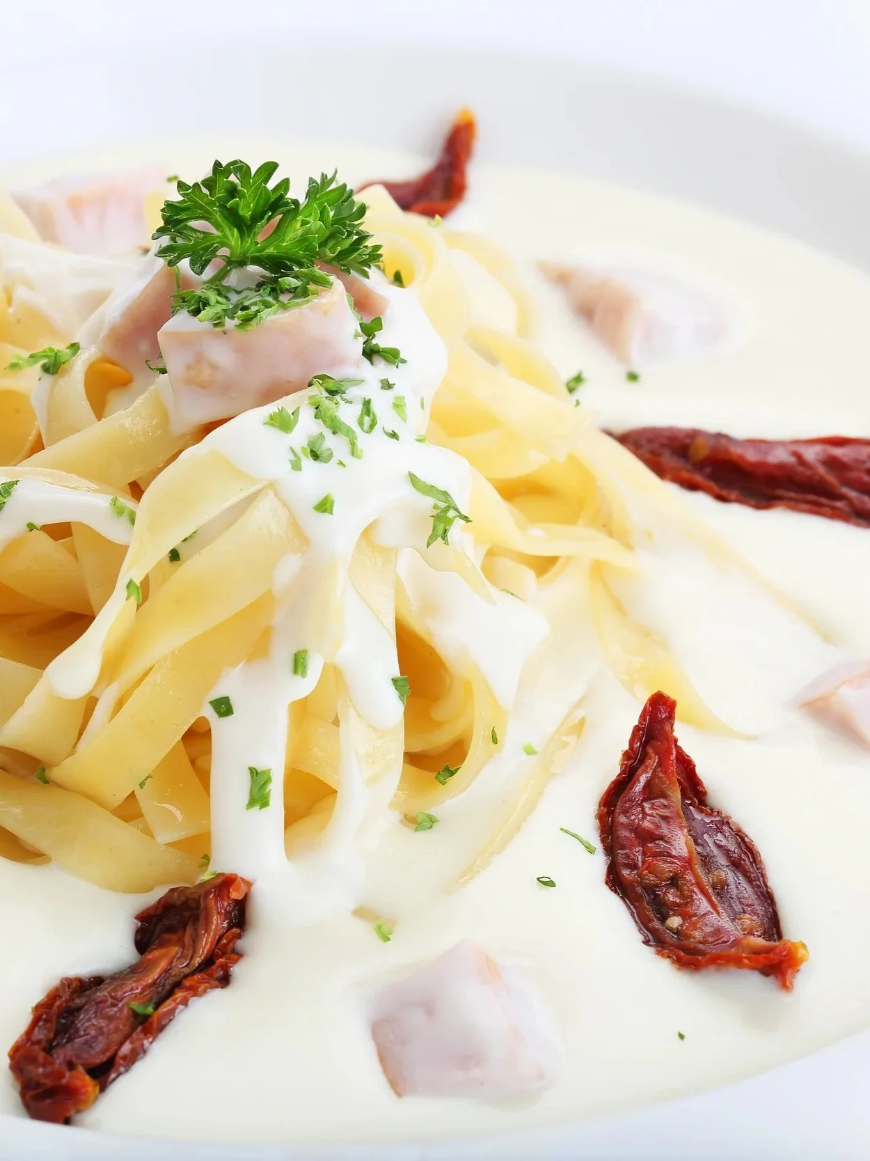 Creamy fettuccine pasta with Alfredo sauce, topped with parsley, sun-dried tomatoes, and diced ham on a white plate.