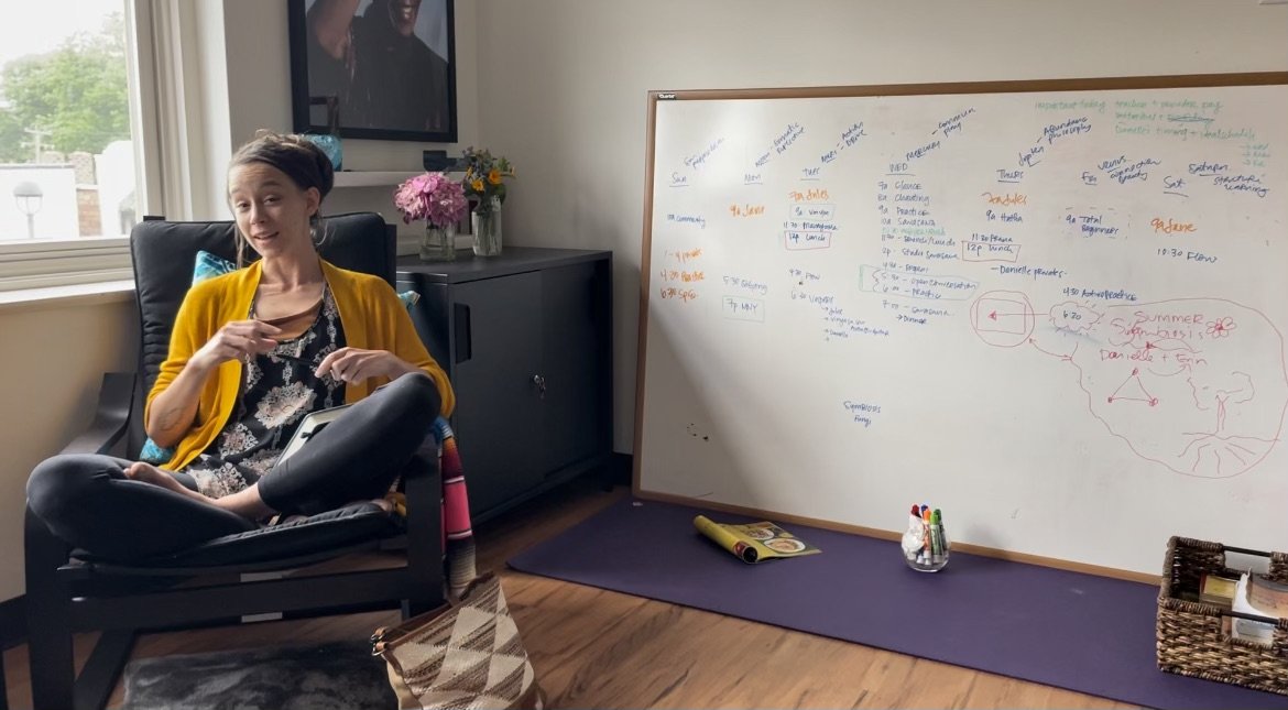Woman sitting in chair next to a whiteboard covered in notes, with cabinets and flowers in the background.