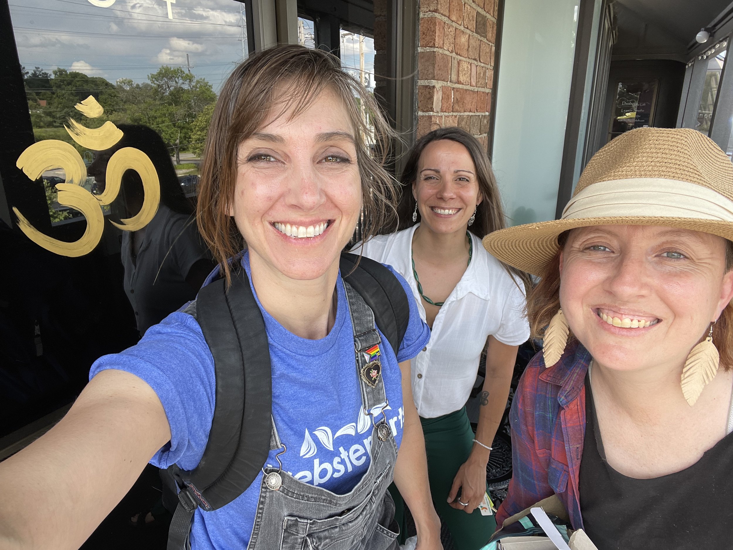 Three women smiling and posing for a selfie, one wearing a backpack, another in a white top, and the third with a straw hat and feather earrings. The background features a decorative 'Om' symbol on a dark window.