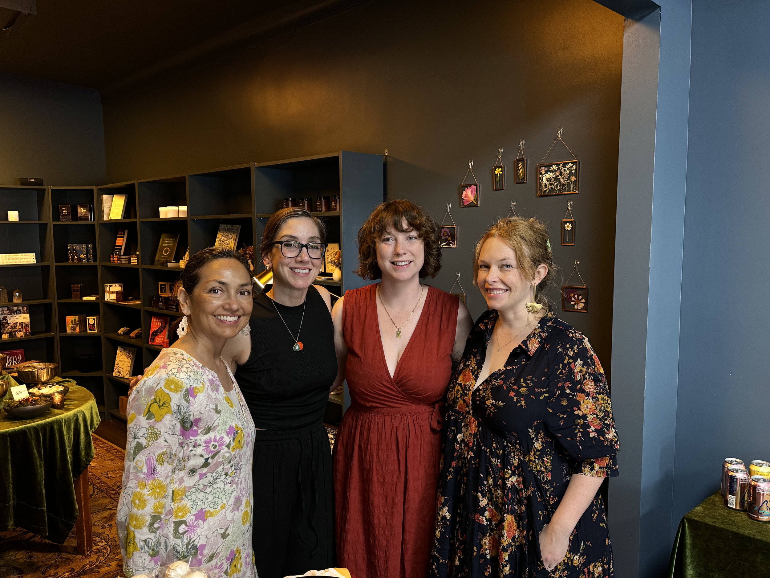Group of four women posing indoors in front of shelves with decorative items and framed art on the wall.