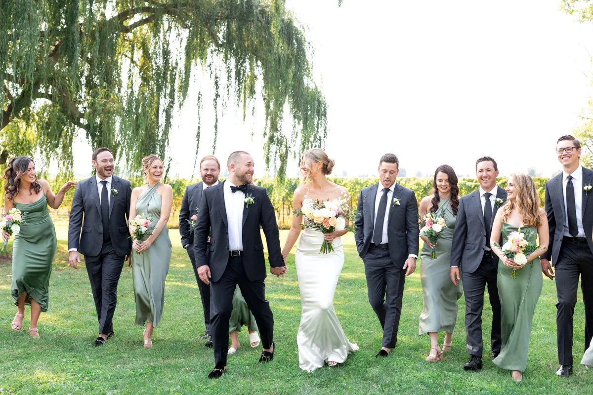 wedding party with bride and groom walking and laughing together with a vineyard and willow tree in the background