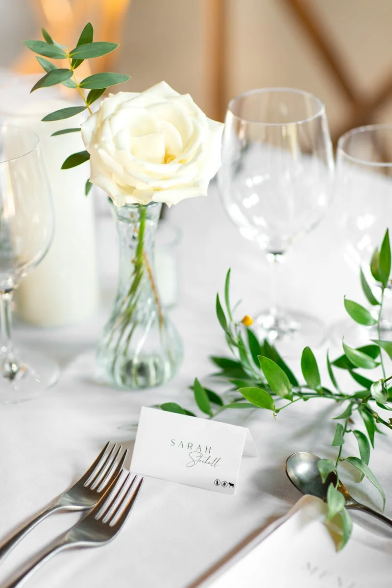 white table setting with white rose in a bud vase and loose greenery with a place card with the guest's name on it