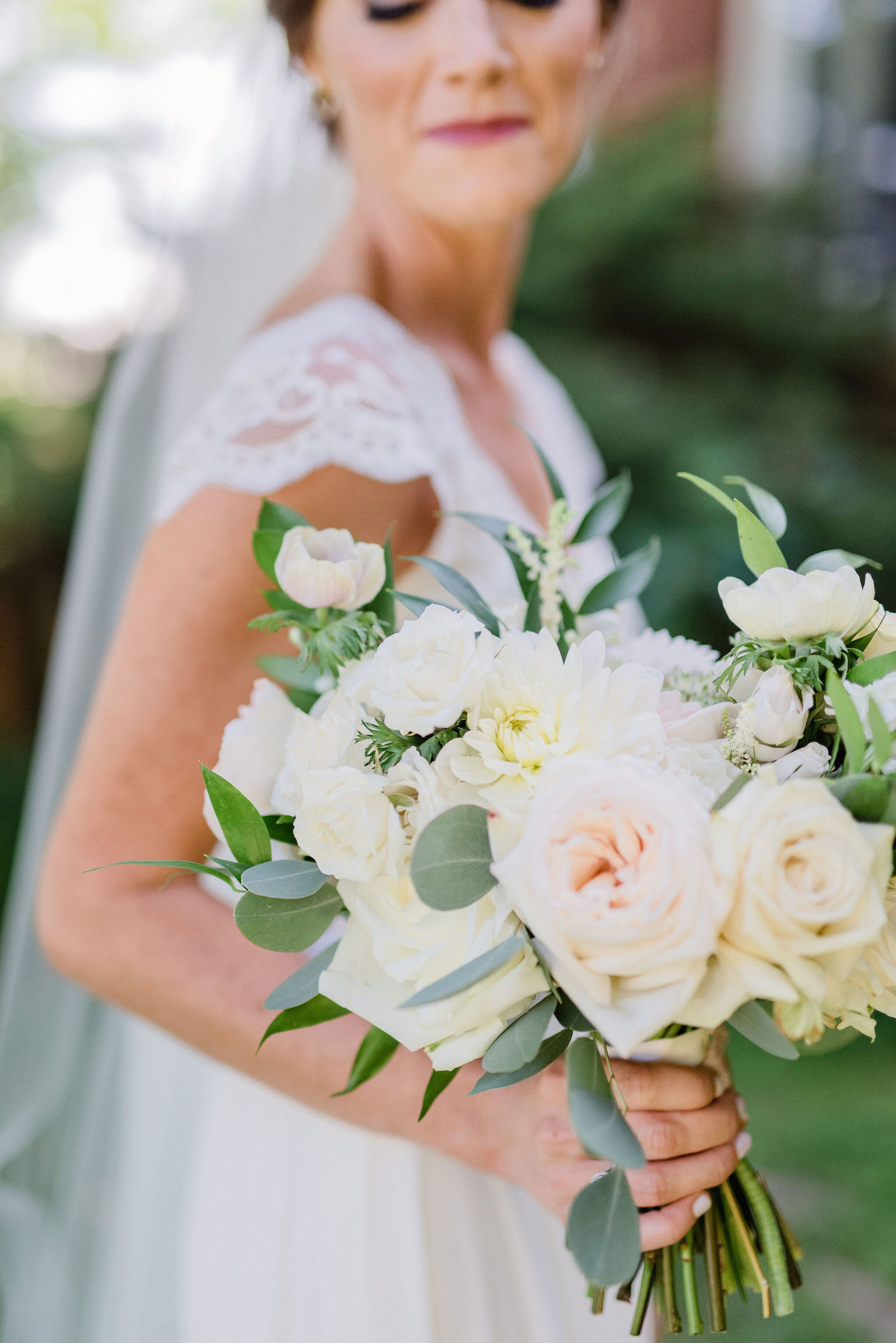 white and blush wedding bouquet with roses and greenery being held by the bride who is glancing down at it