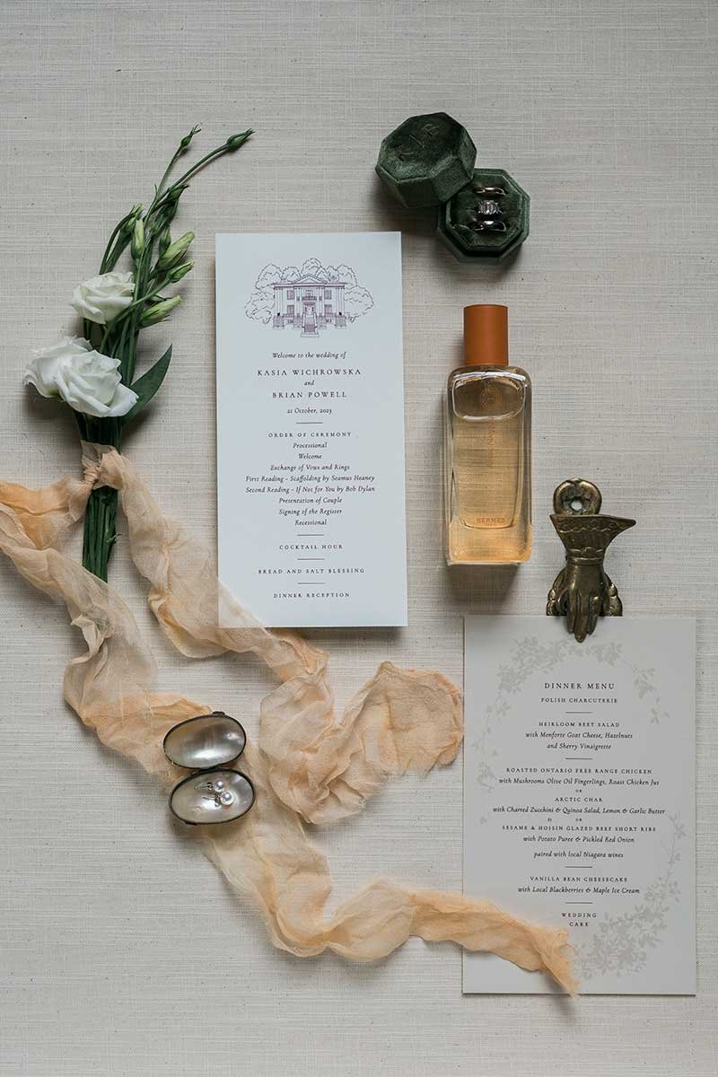  Flatlay of wedding details including invitation, menu, perfume, and jewellery. 