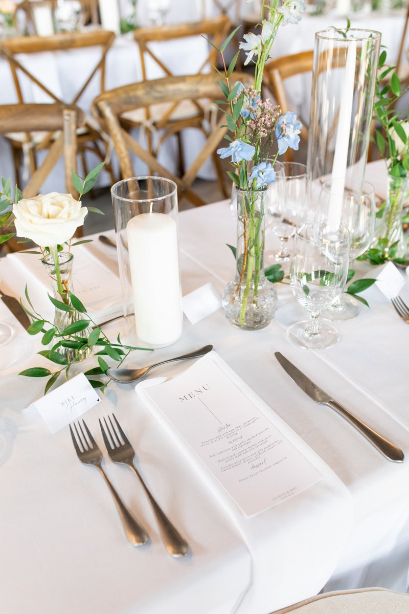 wedding reception table setting with candles and bud vases decorated with greenery, and individual menu card