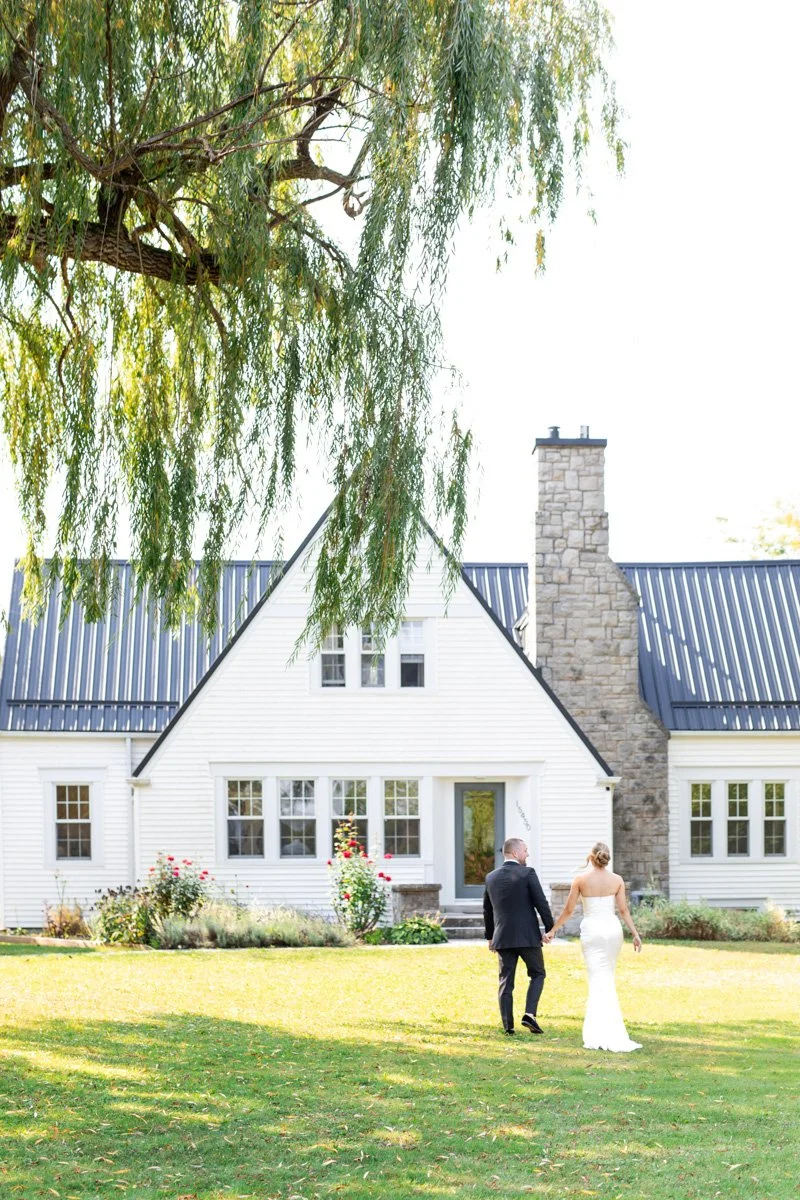 bride and groom walking hand in hand in front of a large white farmhouse