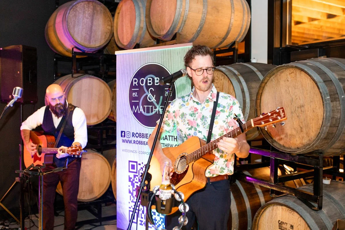 two musicians playing acoustic guitar and singing for a winery wedding reception with wooden wine barrels in the background