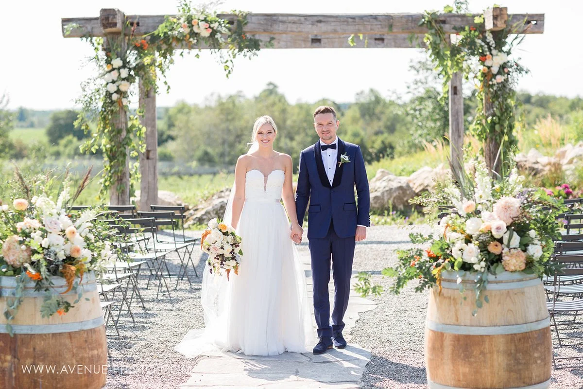 bride and groom standing at an outdoor ceremony site overlooking a vineyard flanked by wine barrels topped with floral arrangements