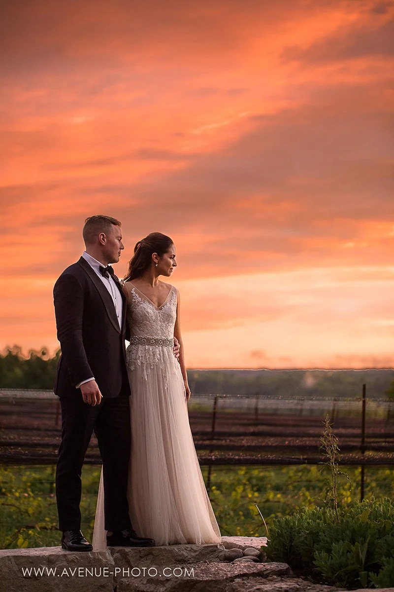 bride and groom standing in a vineyard with a beautiful orange sunset glow behind them