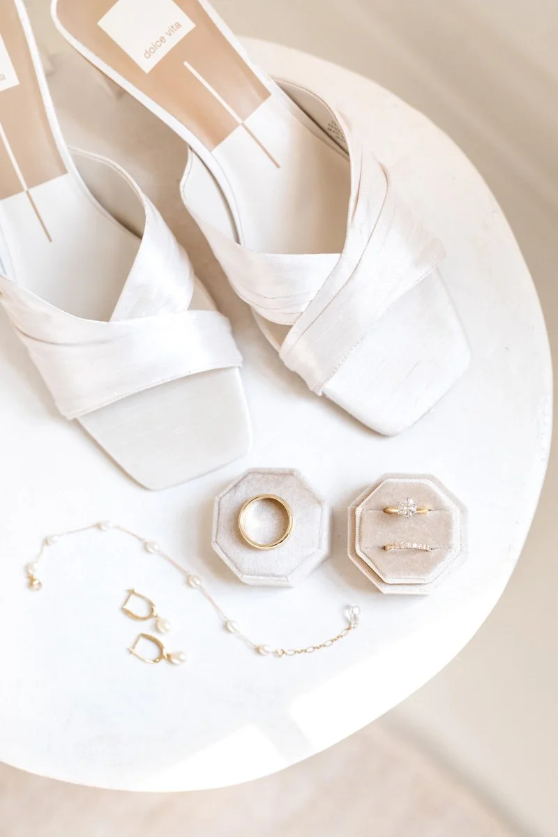 ivory bridal shoes on a marble table with gold jewelry and velvet ring box