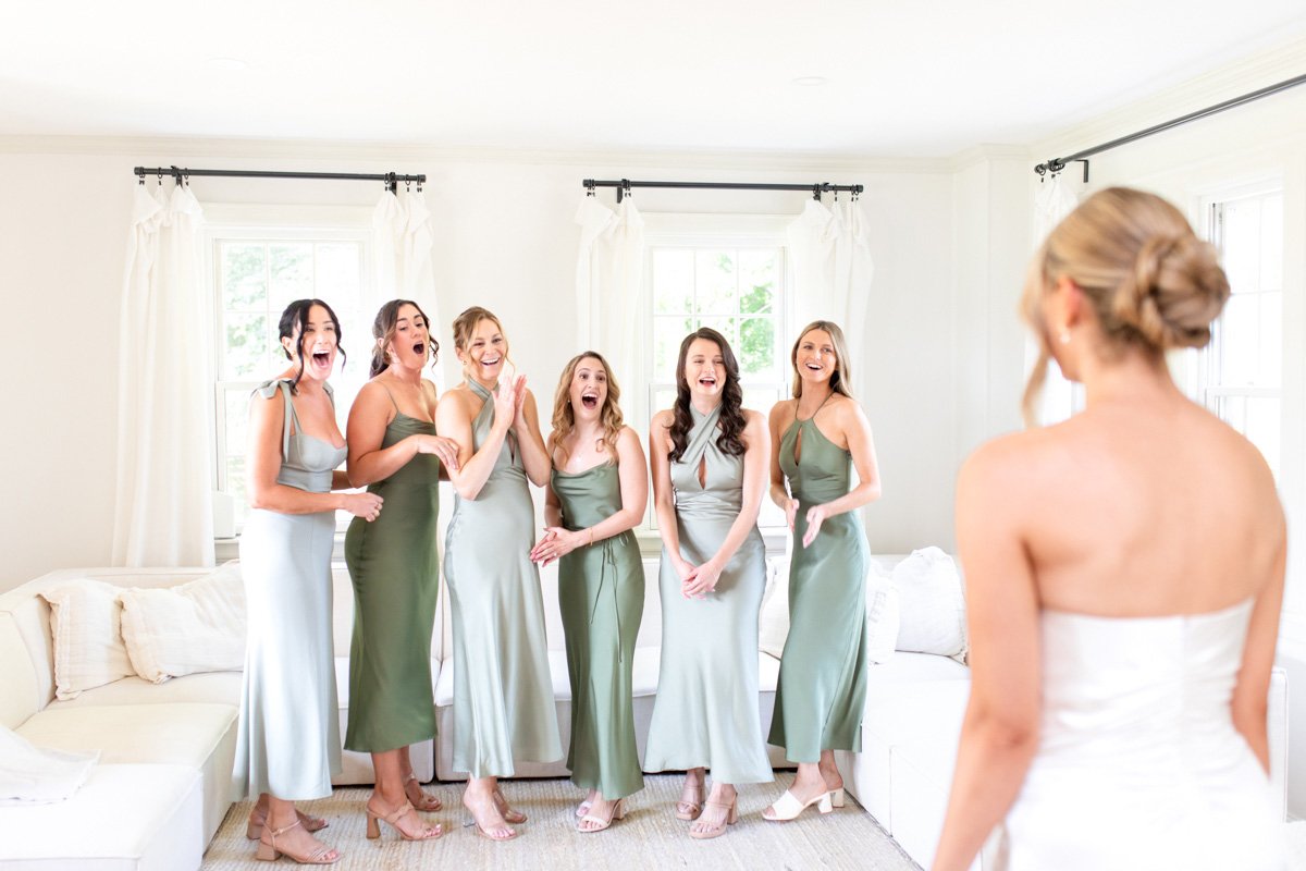 bridesmaids in various sage tone dresses exclaiming in surprise seeing the bride