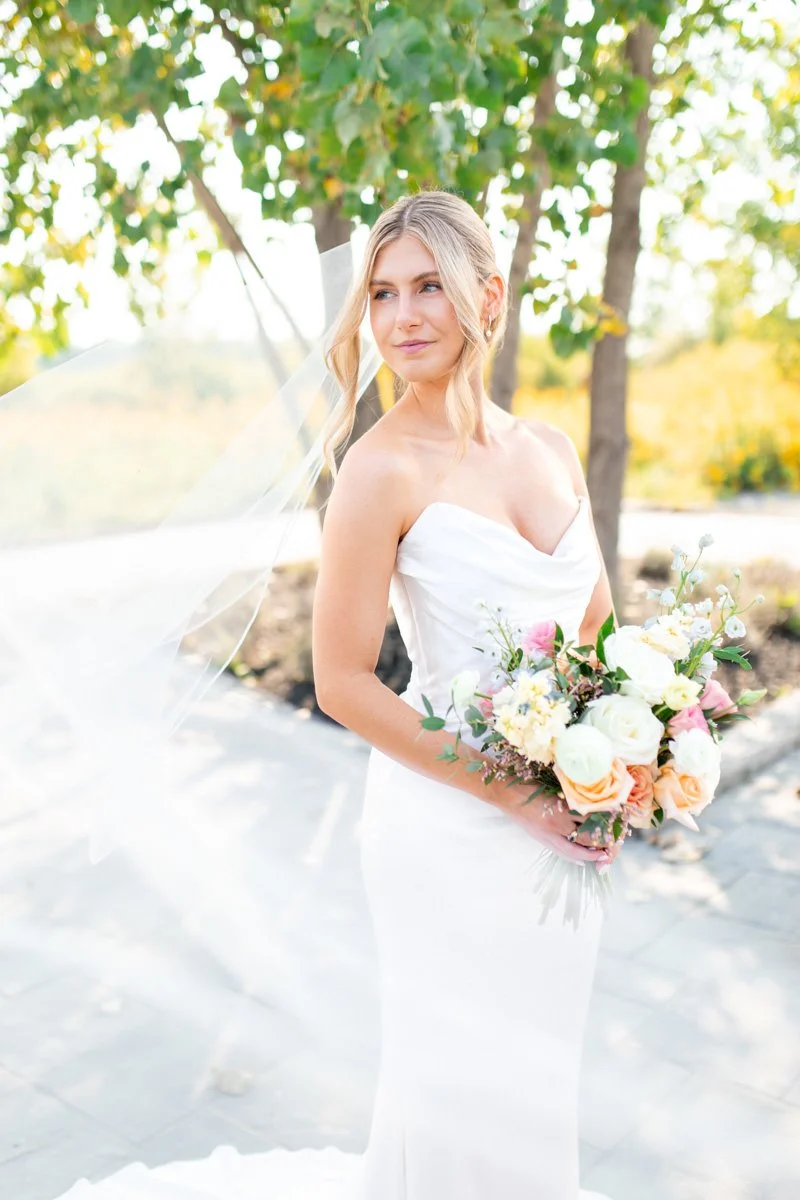 bride with blonde hair in an undo style holding a flower bouquet looking away from the camera while her long veil blows in the wind