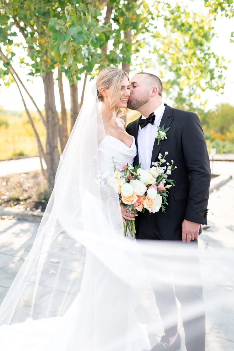 groom wearing a black suit ad bowtie kisses his bride on the cheek while her  long veil blows in the wind