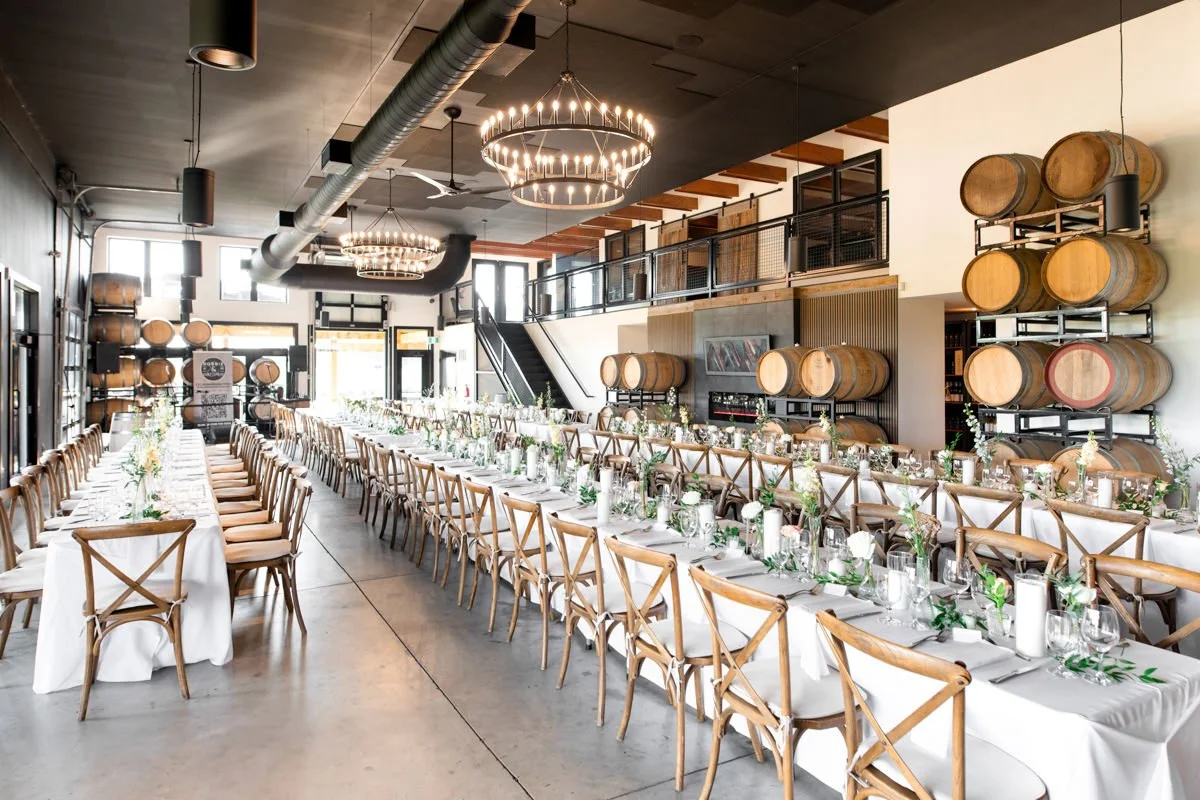 winery wedding reception space amidst wine barrels, long harvest tables with wooden chairs, candles and greenery decorating the lengths of each table