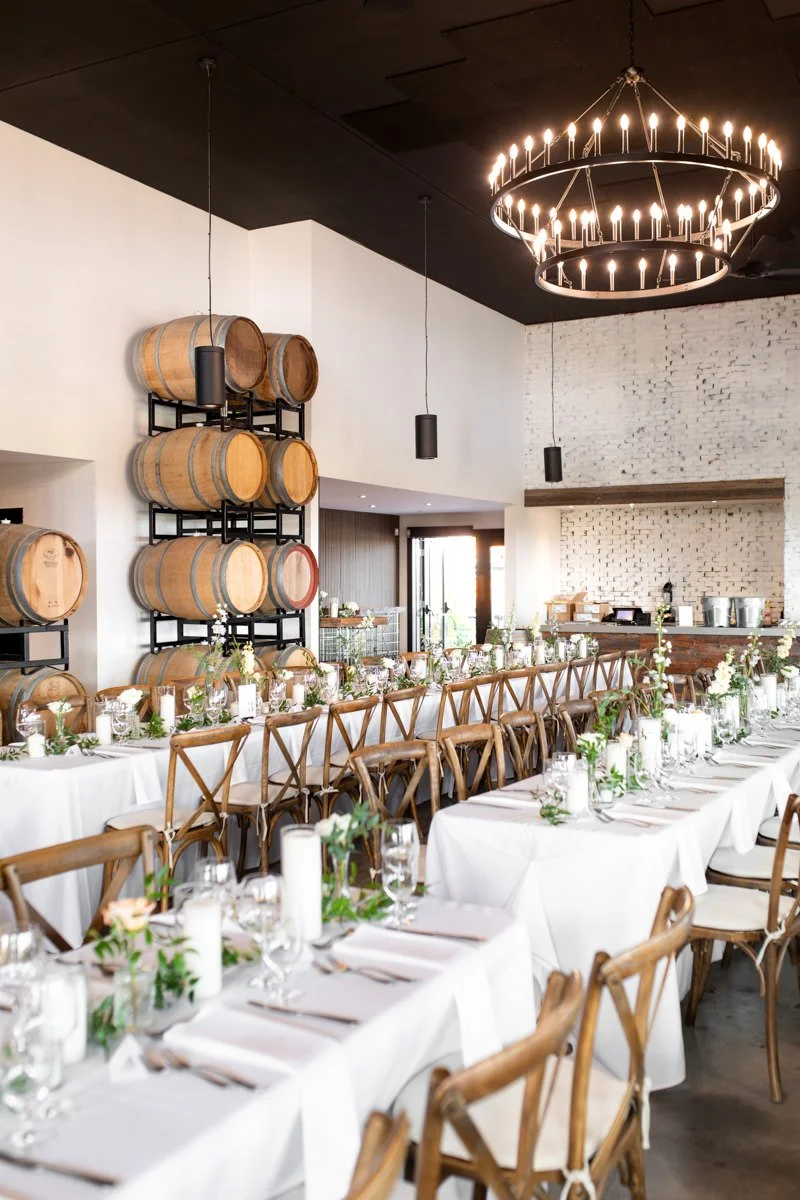 winery wedding reception with long harvest tables decorated in greenery and candles with wooden chairs and wooden wine barrels