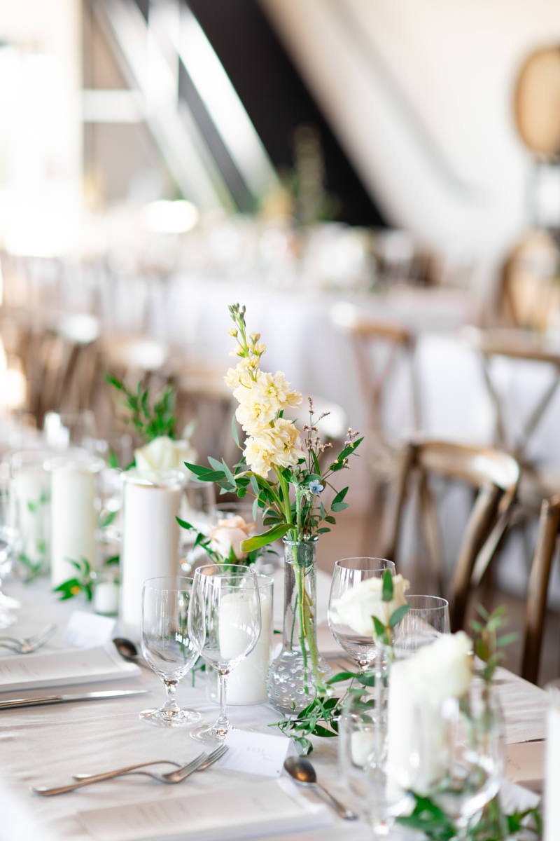 close up of small glass bud vases decorating long harvest wedding reception tables, filled with greenery and small flowers