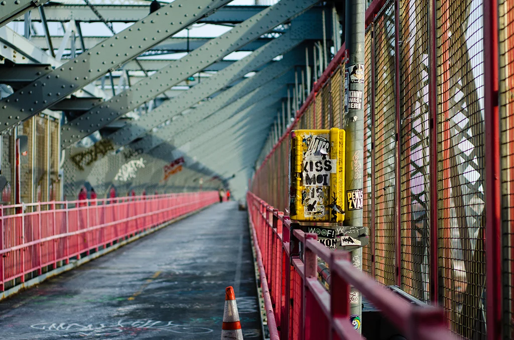 Empty pedestrian bridge with red railings, covered by a metal structure, decorated with graffiti and stickers, with a traffic cone on the wet pavement.