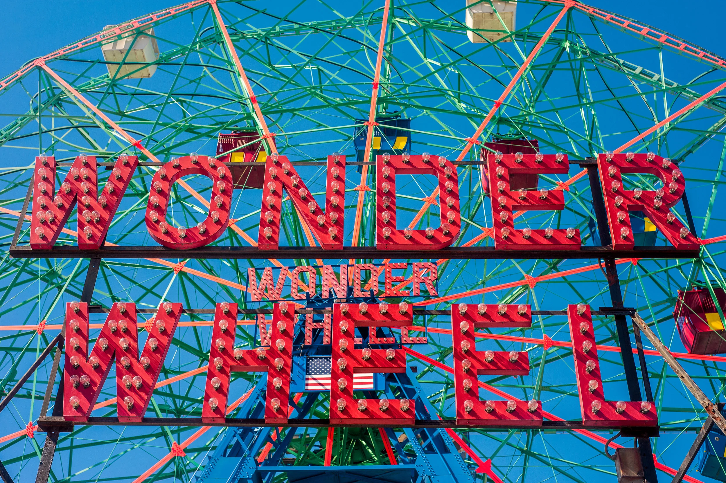 Brightly lit neon sign reading 'Wonder Wheel' in front of a colorful Ferris wheel with a clear blue sky background in Coney Island, Brooklyn, New York.