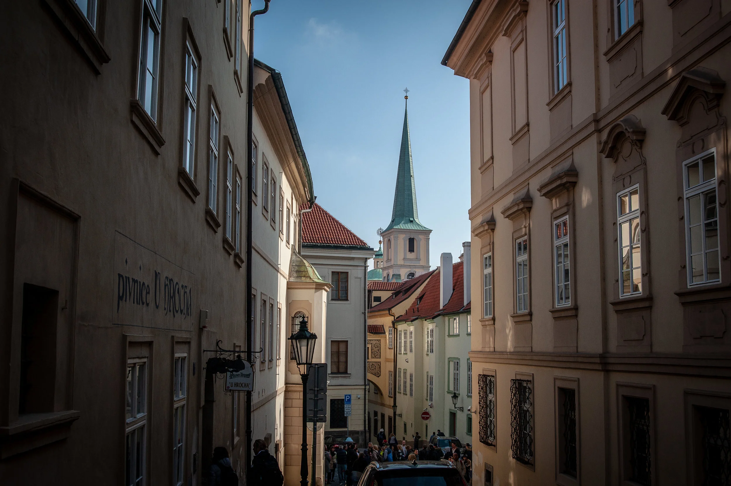 Narrow street in a European city with historic buildings, a church with a tall spire in the background, and people walking on the sidewalk.