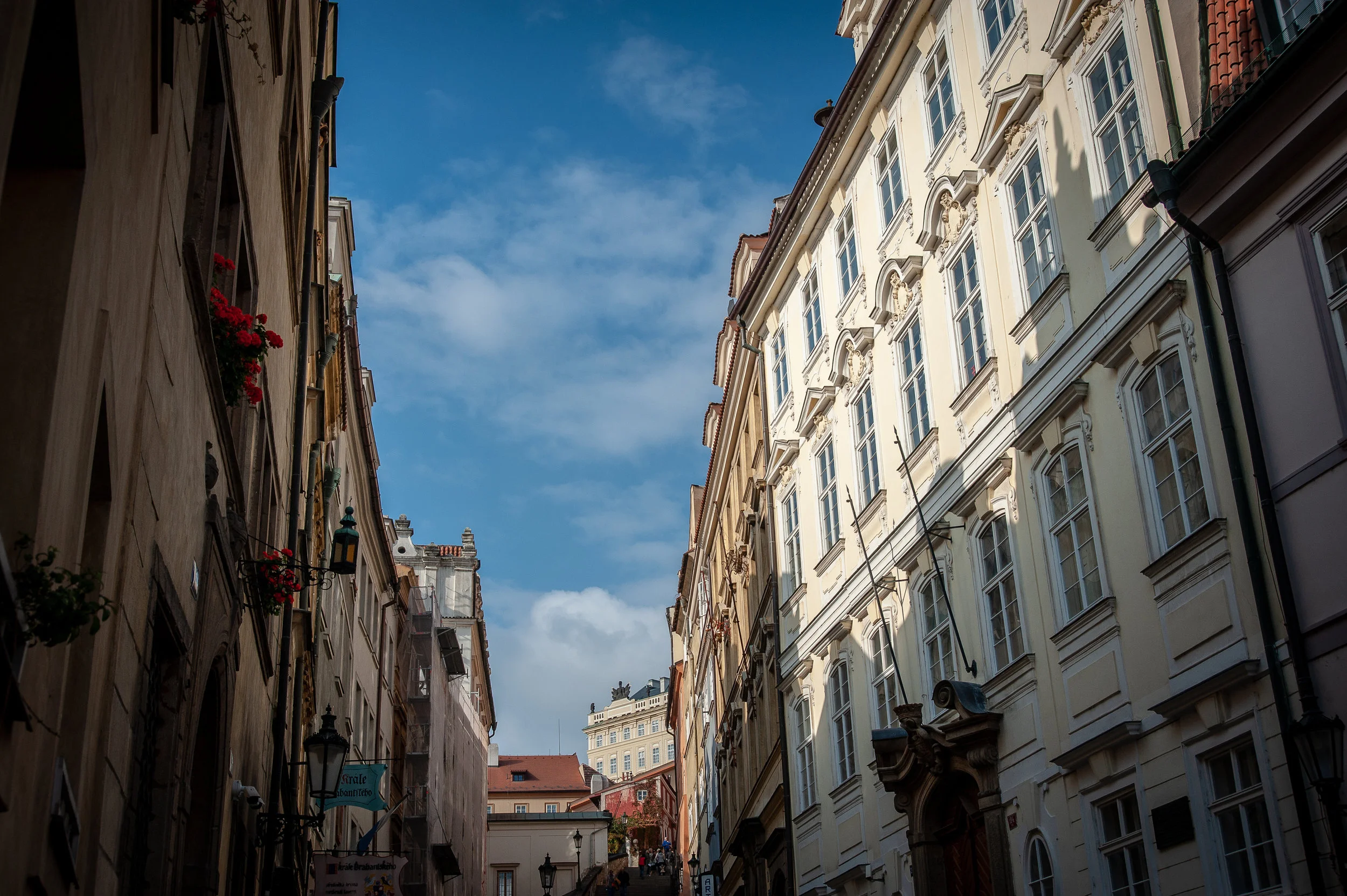 A narrow street lined with historic buildings on both sides, with a clear blue sky overhead and a few clouds.