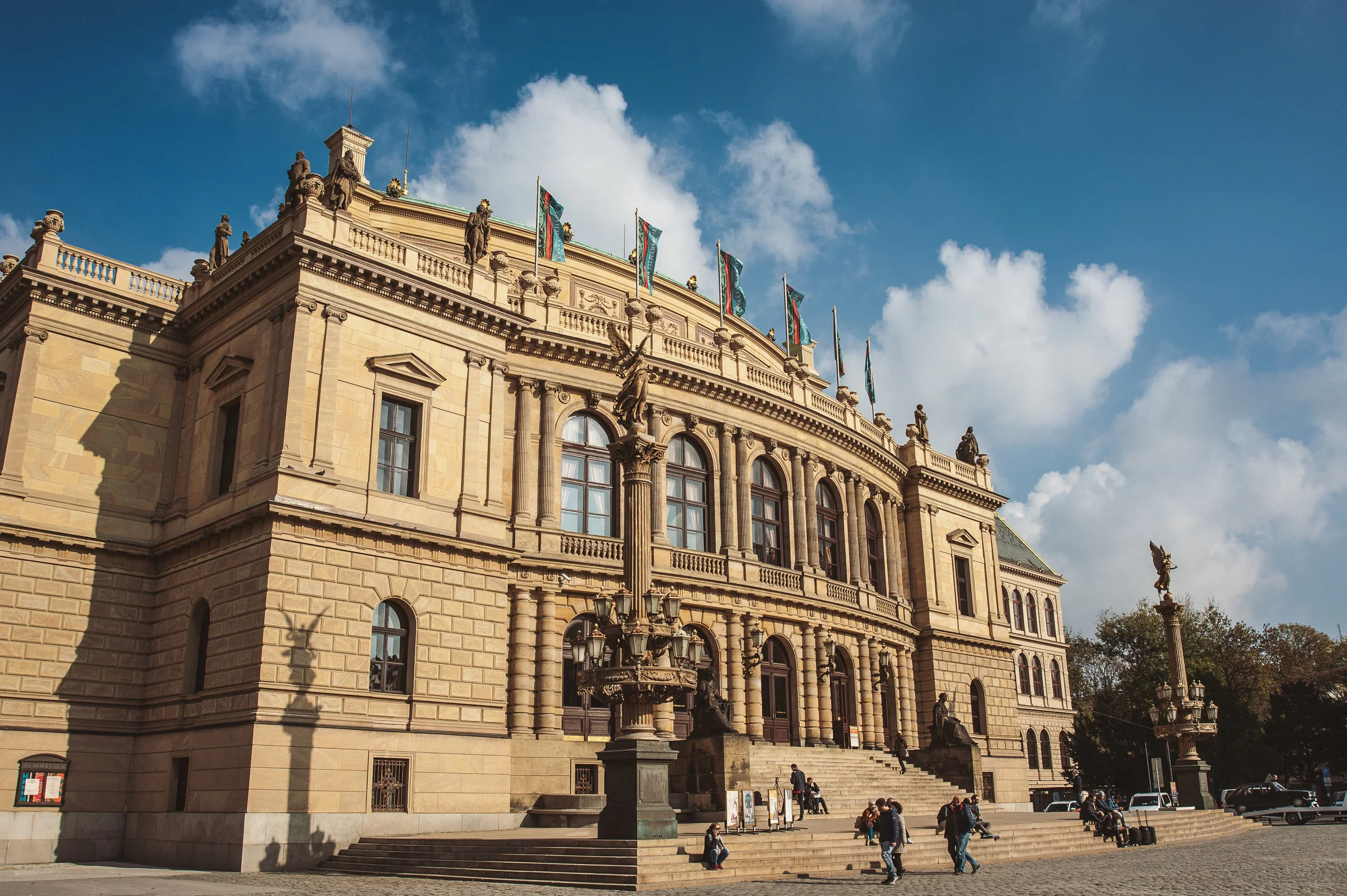 A large historic beige stone building with multiple windows, statues on the roof, and flags flying. People are sitting on the steps in front under a partly cloudy sky.