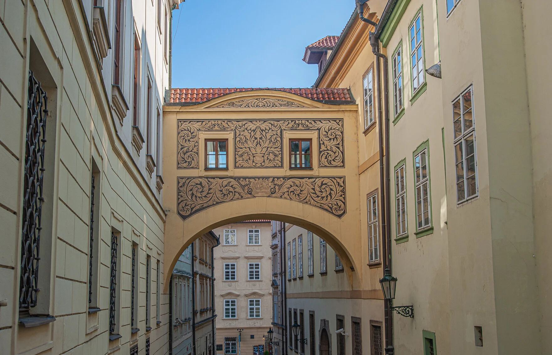 Narrow European street with yellow and cream buildings, decorated archway connecting buildings, and ornate inscription panel above arch, under a blue sky.