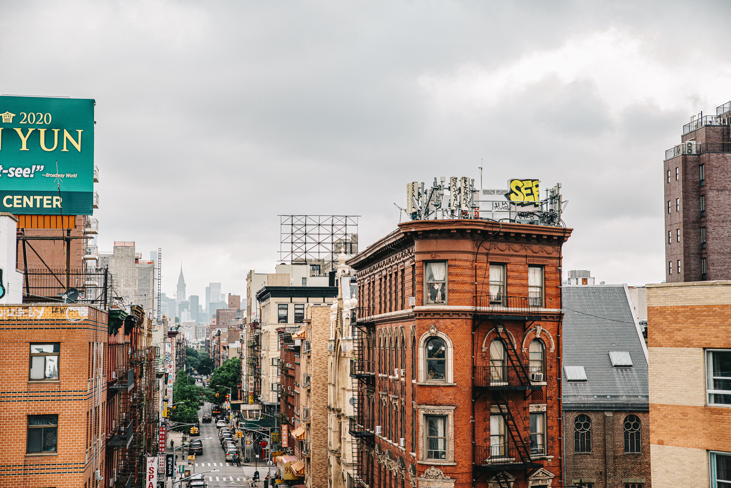 A New York City cityscape view of a street lined with older brick buildings, some with fire escape ladders, and a distant skyline with tall skyscrapers under cloudy skies.