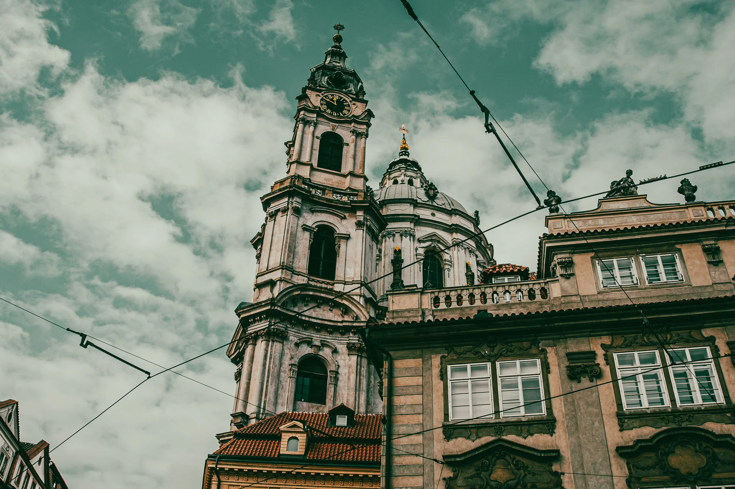 View of a historic church in Prague with a tall clock tower surrounded by buildings and power lines against a cloudy sky.