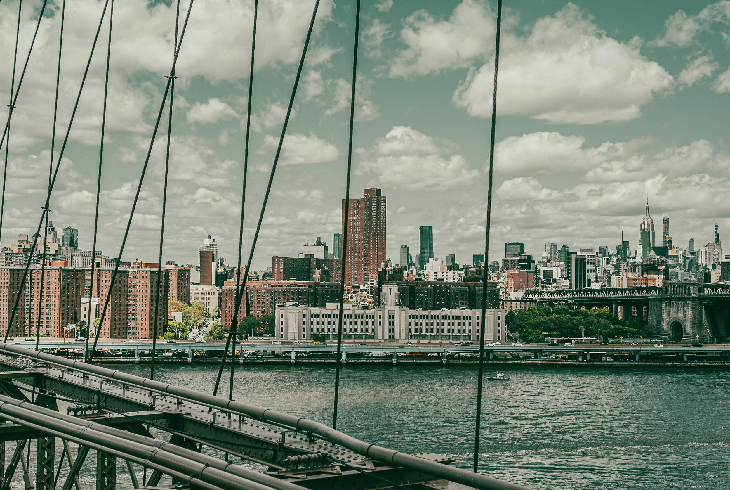 The New York City skyline with tall buildings viewed from a bridge over the East River, with cables and structural elements of the Brooklyn Bridge in the foreground and partly cloudy sky overhead.