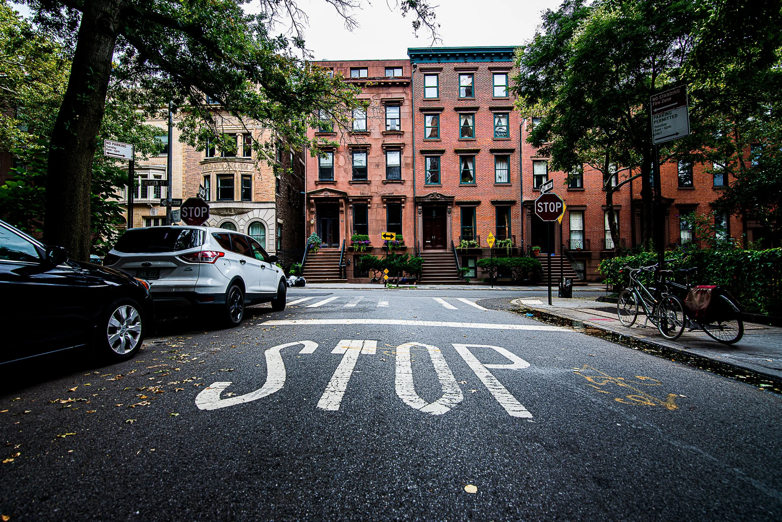 A city street in Brooklyn Heights, New York with a large stop sign painted on the road, parked cars, bicycles, trees, and brick residential buildings in the background.