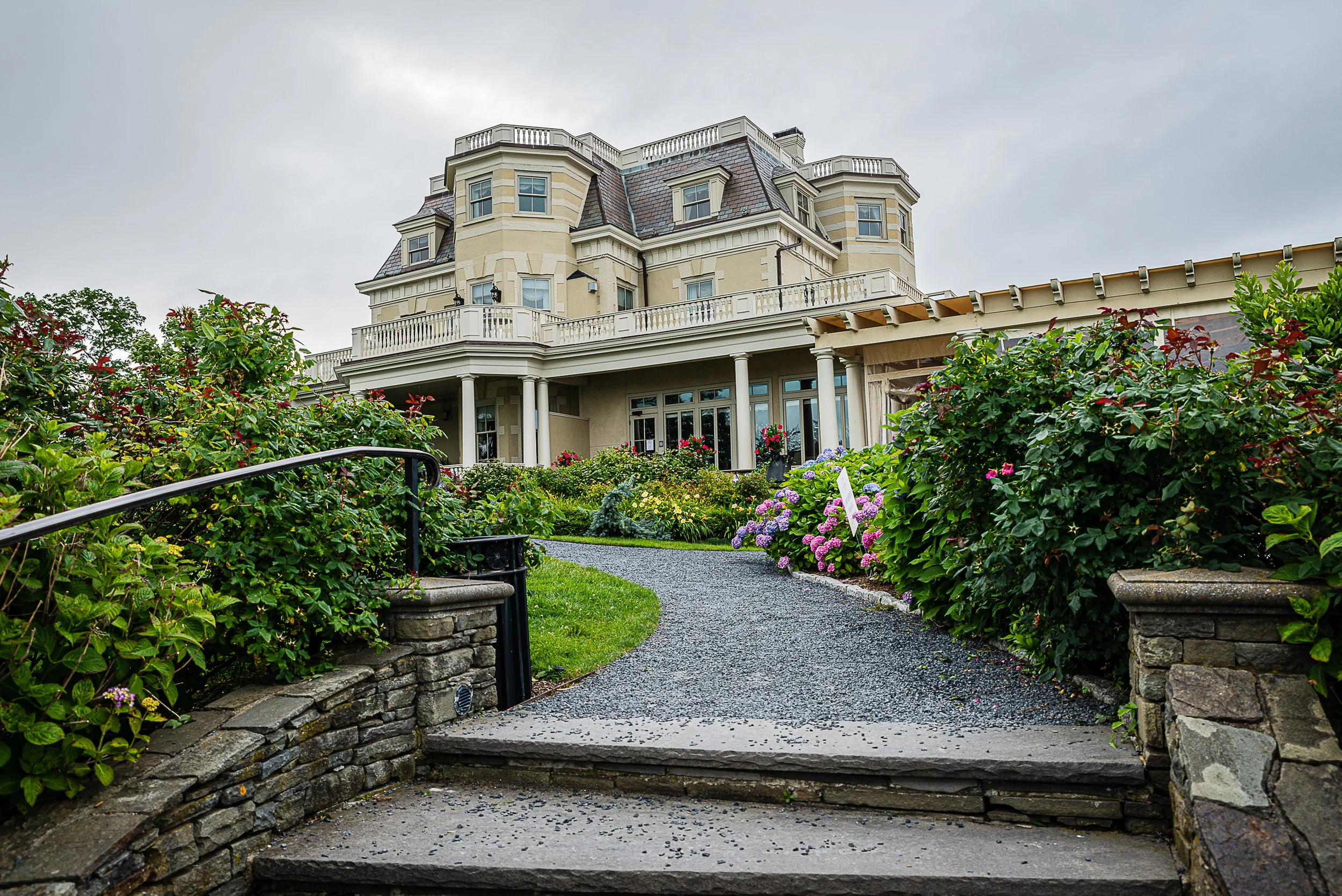 Large, elegant mansion in Newport, Rhode Island with multiple levels and balconies, surrounded by a lush, well-maintained garden with colorful flowers and a winding pathway leading to the entrance.