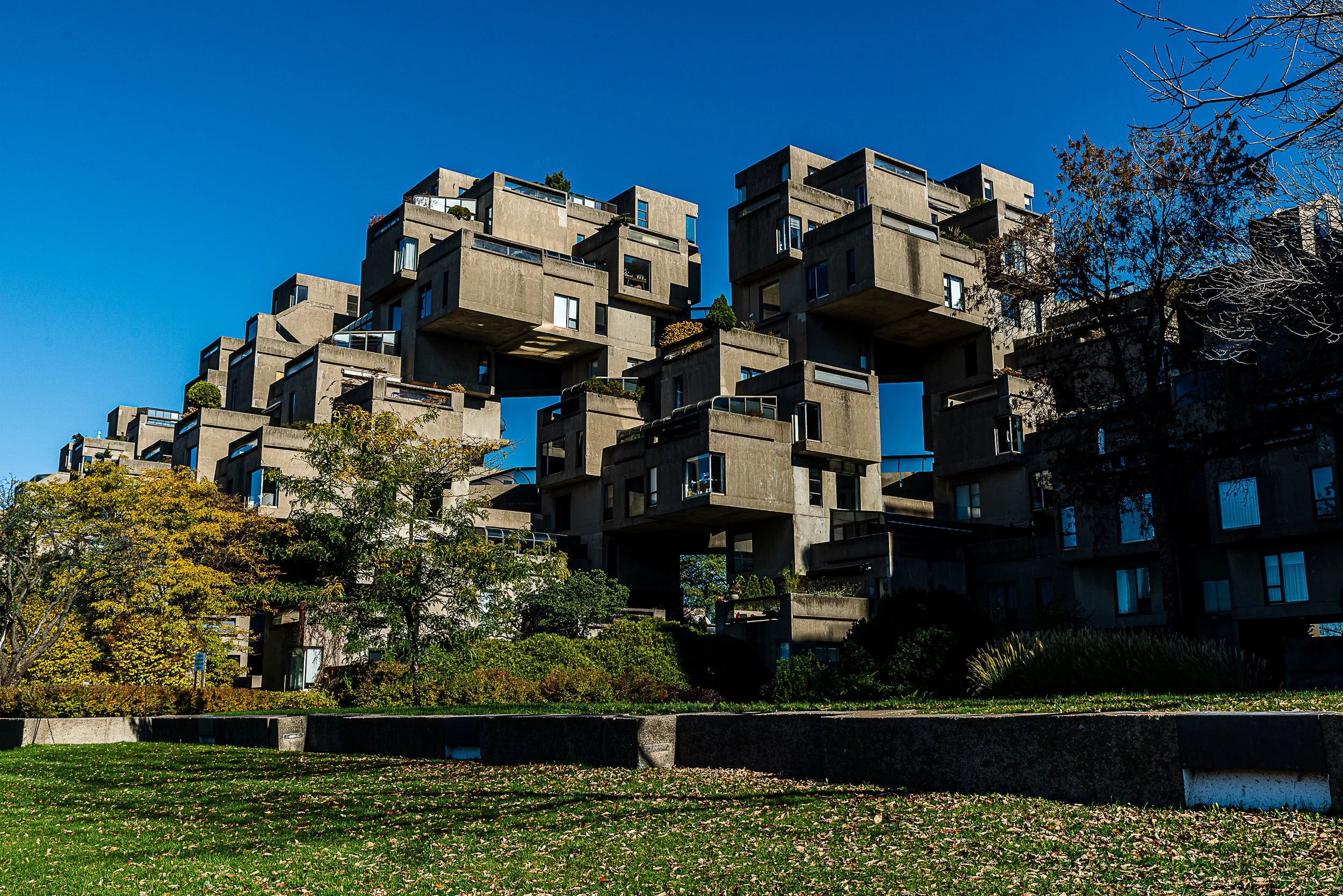 Unique concrete apartment building named Habitat 67 in Montreal, Canada with multiple box-like structures, surrounded by trees and green space, under a clear blue sky.